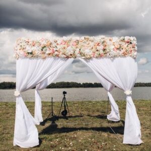 beautiful wedding arch with flowers.view over white wedding arch with flowers on the top and rose petals scattered on the grass. situated by the river on cloudy day.