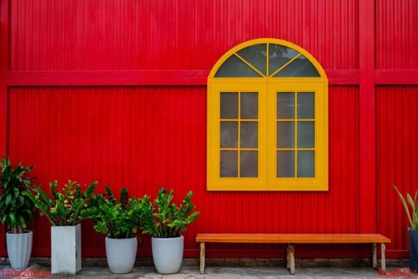 a large yellow window, flower pots with plants and a bench against a red metal wall on a city street .