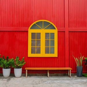 a large yellow window, flower pots with plants and a bench against a red metal wall on a city street .