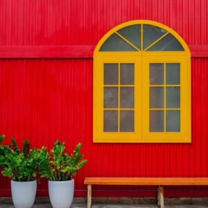 a large yellow window, flower pots with plants and a bench against a red metal wall on a city street .