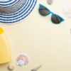 beach hat, sunglasses and sunscreen are among the seashells on a beige surface